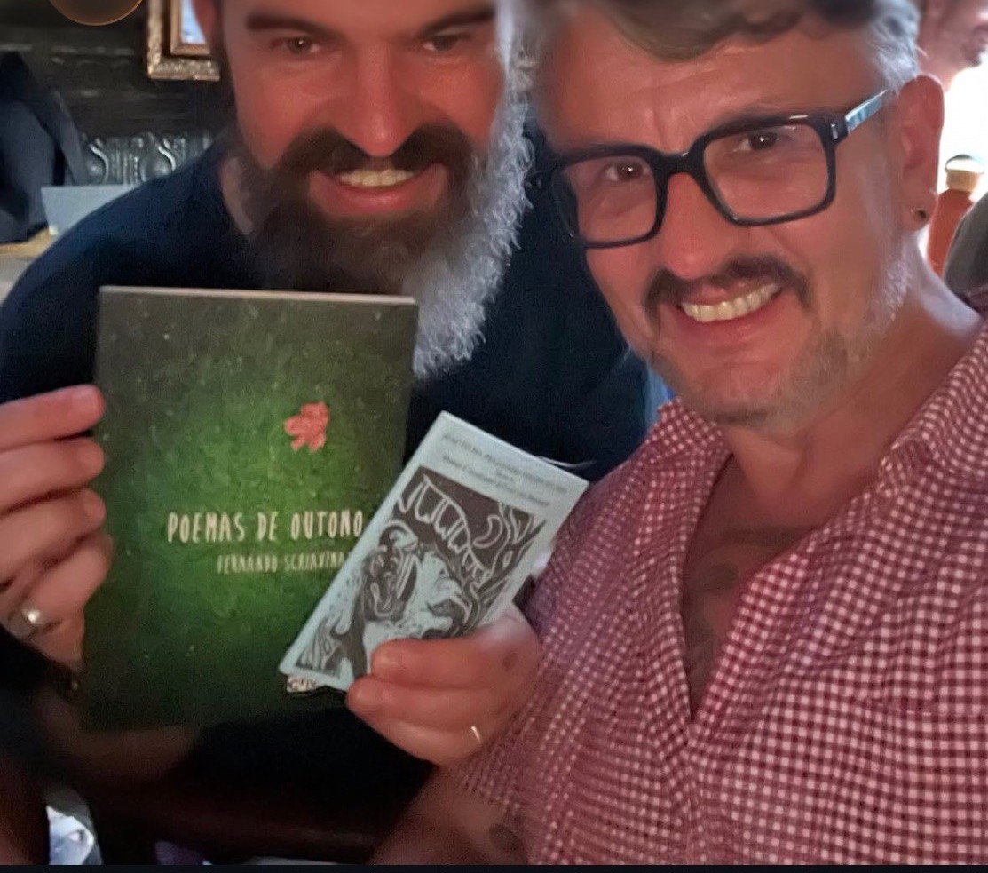 Two smiling guests hold copies of “Poemas de Outono” and another book on the night of the sarau.