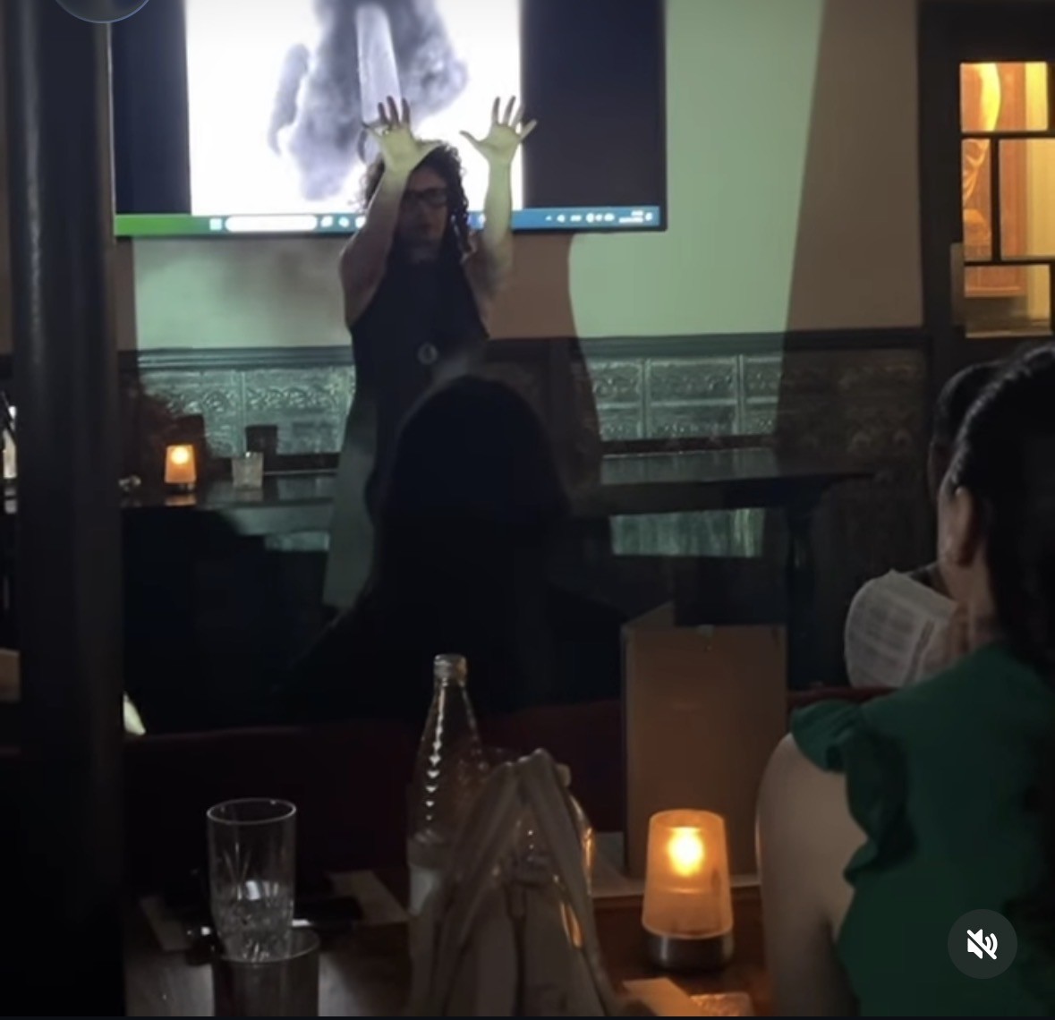 A performance with raised hands in front of a projection, seen across candlelit tables.
