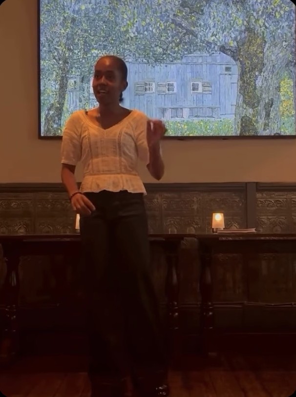 Aleida Tavares standing and speaking to the room, in front of a painting and a table set with candles.